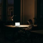 A lone man working on a laptop under dim lighting in a quiet office at night.
